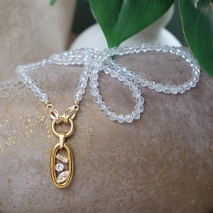 May include: A necklace with clear, faceted beads and a gold-coloured pendant. The pendant has an oval frame with three clear stones. The necklace is displayed on a textured, neutral-toned surface. The necklace's length is approximately 45 cm.