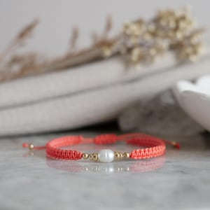 May include: A coral-colored cord bracelet featuring a central white freshwater pearl flanked by two small gold-toned metallic beads on each side. The bracelet is constructed with a woven macrame knot pattern and includes adjustable sliding cord ends.