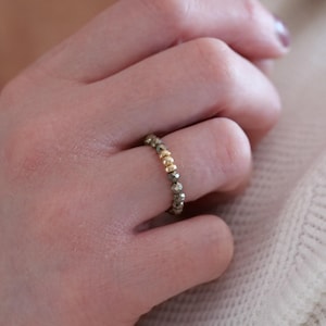 Könnte beinhalten: Ein Perlenring mit kleinen, facettierten Pyritsteinen und goldfarbenen Perlen. Der Ring wird an einem Finger getragen und zeigt den Kontrast zwischen dem metallischen Gold und dem strukturierten Pyrit. Das Design des Rings ist schlicht, aber elegant und für den Alltag geeignet.