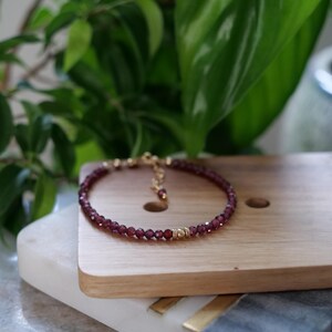 May include: A delicate bracelet featuring small, faceted, deep red beads strung together with gold-colored accents. The bracelet is displayed on a wooden surface, with green foliage in the background.