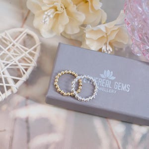 Könnte beinhalten: Zwei Perlenringe, einer in Gold und einer in Silber, werden auf einer grauen Schachtel mit dem Text "Little Real Gems Jewellery" präsentiert.