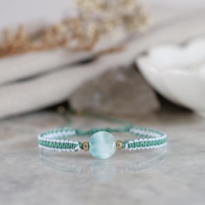 Peut inclure: Un bracelet en macramé avec une perle centrale ronde en pierre bleu clair, encadrée par deux petites entretoises en métal doré. Le bracelet est composé d'un cordon tressé vert sauge et blanc, formant un motif texturé autour du poignet.