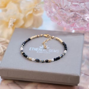May include: A delicate bracelet with black, white, and gold beads, resting on a gray box. The bracelet is made of small, round beads and is connected by a gold chain. The box has the text "ETHEREAL GEMS JEWELLERY".
