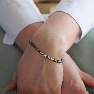 May include: A beaded bracelet featuring small, dark gray, faceted hematite beads alternating with tiny, polished gold-tone metal spacer beads. The bracelet is strung on a thin, flexible cord and sits snugly around the wrist, showcasing a simple design.