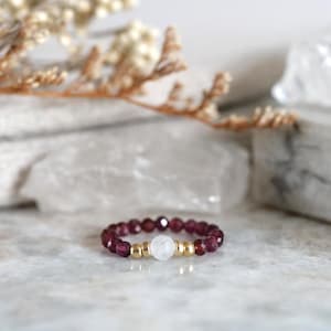 May include: A beaded ring featuring a series of small, faceted deep red garnet beads arranged in a circular band. The centre of the ring displays a single round, smooth white stone bead flanked by two small, polished gold-toned metallic beads.
