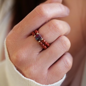 May include: A close-up of a hand wearing two stacked rings. The rings are made of small red beads with a gold accent. The top ring features a square-shaped stone with a red, blue, and green pattern.