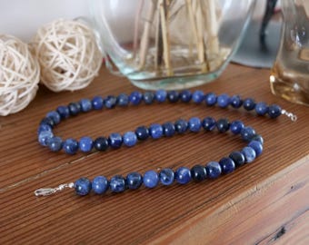 Puede incluir: Un collar de cuentas compuesto por piedras de sodalita redondas y pulidas en diversos tonos de azul profundo y azul marino. Las cuentas están ensartadas en una sola hebra, terminada con un cierre de mosquetón de metal plateado en cada extremo.