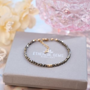 Peut inclure: Un bracelet en pierres grises avec des perles dorées et un fermoir. Le bracelet est présenté dans une boîte grise avec le texte "ETHEREAL GEMS JEWELLERY".