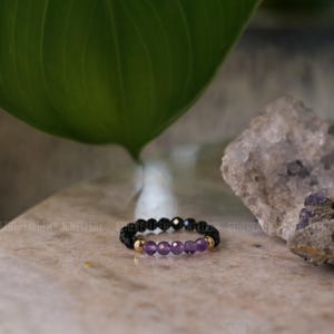 May include: A beaded ring with black, gold, and purple gemstones. The ring has a band of black beads, with gold accents and a row of purple amethyst stones in the centre. The ring is displayed on a light-coloured surface.
