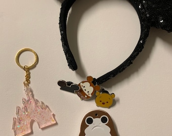 Könnte beinhalten: Ein Set an Disney-Accessoires, bestehend aus einem schwarzen und roten, mit Pailletten besetzten Minnie Mouse-Haarreif, einem pinkfarbenen Glitzer-Schlüsselanhänger mit einem Schloss und einem braunen und weißen Schlüsselanhänger mit einem Star Wars-Charakter und einem rosa Knopf.