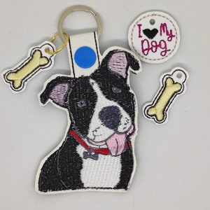 Puede incluir: Un llavero de perro bordado en blanco y negro con un collar rojo y una lengua rosa que sobresale. El llavero también incluye un pequeño amuleto de hueso y un amuleto redondo que dice "Amo a mi perro" con un corazón rosa.