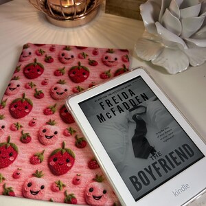 May include: A white Kindle e-reader with a clear case is displaying the cover of the book "The Boyfriend" by Freida McFadden. The e-reader is resting on a pink and white fabric case with a strawberry pattern.