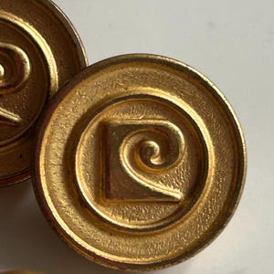 Peut inclure: Un ensemble de boutons ronds en métal doré arborant un logo en forme de lettre P stylisée en relief au centre. Chaque bouton présente un fond texturé en creux à l'intérieur d'un bord circulaire lisse, avec une finition métallique classique.