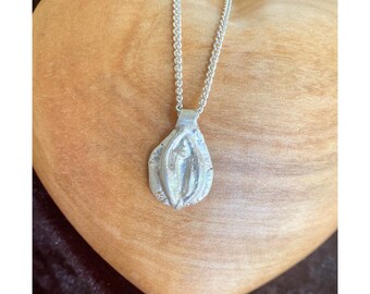 Könnte beinhalten: Eine silberne Halskette mit Anhänger, die ein stilisiertes Vulva-Design aufweist. Der Anhänger hängt an einer filigranen Kette. Die Halskette liegt auf einem herzförmigen Holzgrund. Ein einzigartiges Schmuckstück.