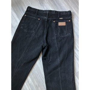 May include: Black denim jeans featuring a high-rise waist, belt loops, and two back pockets. The right back pocket displays a brown leather patch with the Wrangler brand name, and both pockets feature a stitched W design in black thread.