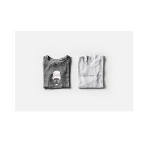 May include: Two folded t-shirts, one gray and one white. The gray t-shirt has a white graphic of a woman with a bun and the text "Folklorico Hair". The white t-shirt has the text "Folklorico Hair" in black.
