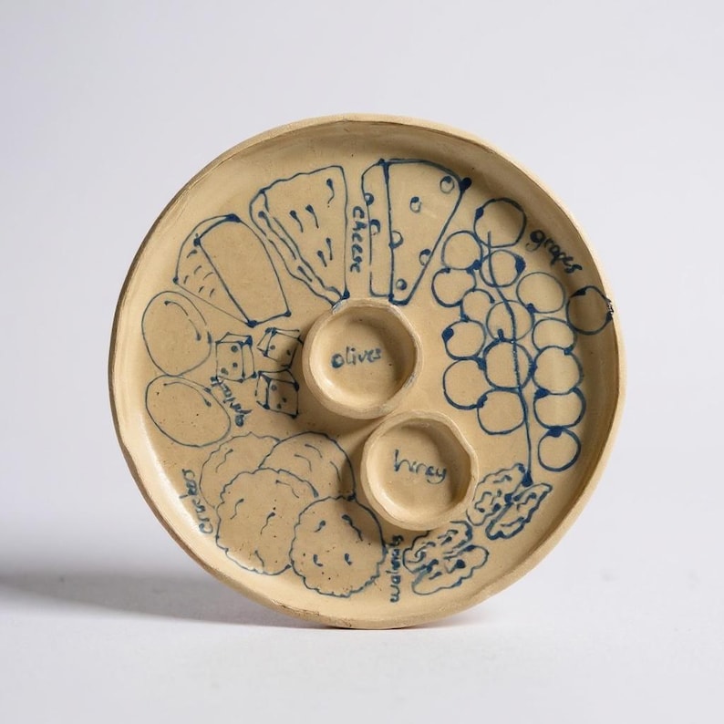 May include: A beige ceramic serving platter with blue hand-drawn illustrations of cheese, grapes, olives, honey, walnuts, and crackers. The platter has two small circular wells for dips.