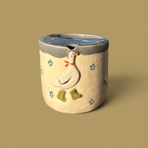 Può includere: Una tazza cilindrica in ceramica con corpo color crema e bordo grigio. L'esterno presenta una figura di oca bianca in rilievo con un fazzoletto rosso al collo e stivali verdi, circondata da piccoli motivi floreali blu dipinti.