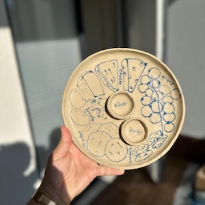 May include: A round, beige ceramic serving platter with blue hand-drawn illustrations of food items. The platter includes sections for cheese, grapes, olives, honey, crackers, and walnuts. The platter is held by a person.