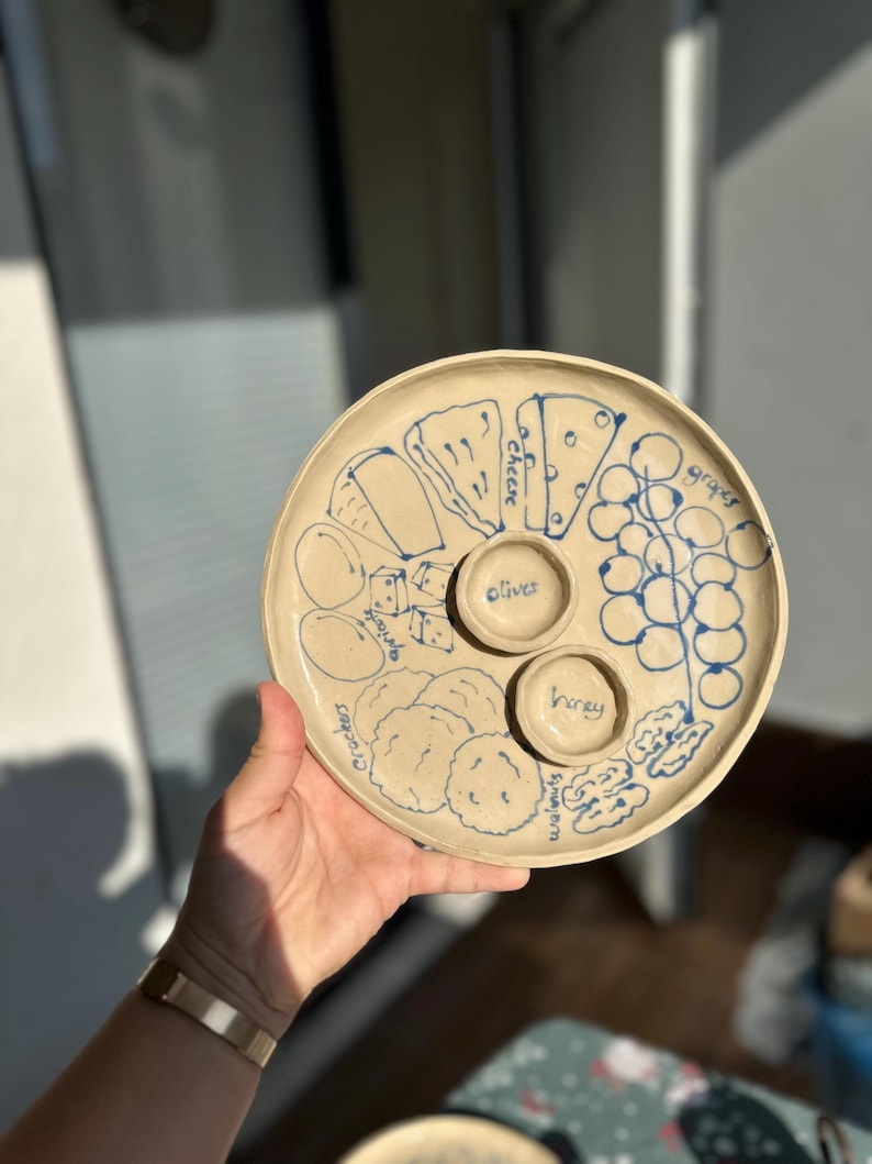 May include: A round, beige ceramic serving platter with blue hand-drawn illustrations of food items. The platter includes sections for cheese, grapes, olives, honey, crackers, and walnuts. The platter is held by a person.