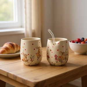 Może przedstawiać: Dwa kremowe ceramiczne kubki z fantazyjnym wzorem królika i kwiatów. Jeden kubek ma zakrzywioną szklaną słomkę. Kubki są ozdobione czerwonymi kropkami i stoją na drewnianym stole. Na stole znajdują się również rogalik i miska jagód.
