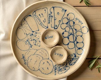Pode incluir: Um prato de servir redondo em cerâmica cor de creme com ilustrações azuis desenhadas à mão. O prato inclui etiquetas para queijo, uvas, azeitonas, mel, bolachas e alperces, com duas pequenas taças integradas no centro.
