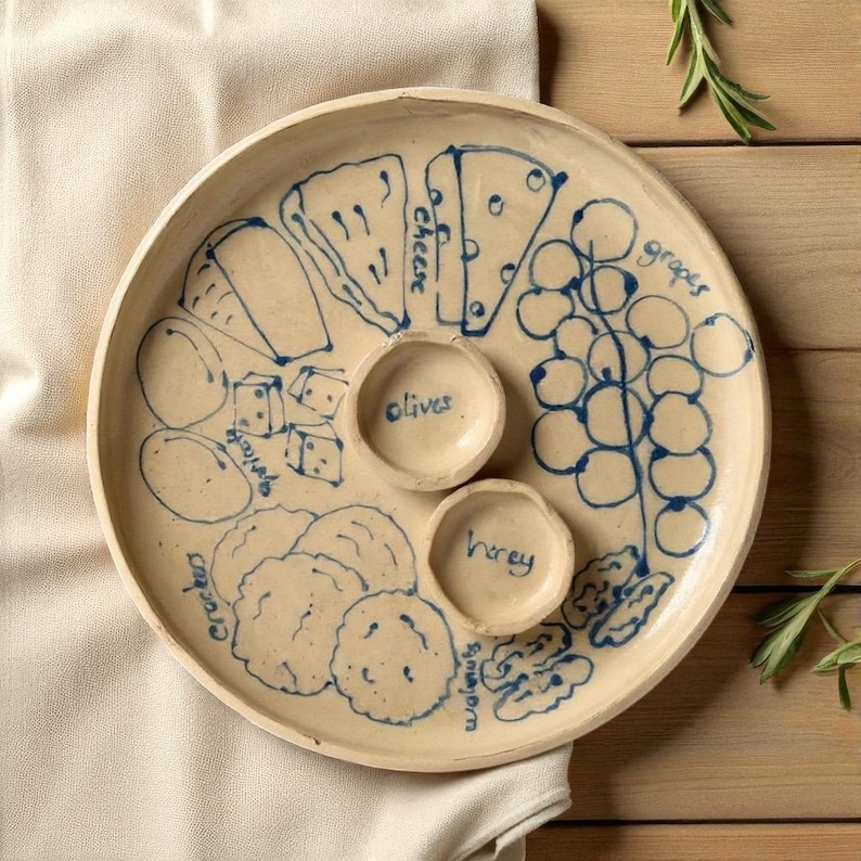 May include: A round, off-white ceramic serving platter with blue illustrations of cheese, grapes, crackers, olives, honey, and walnuts. The platter has two small circular bowls for olives and honey. The words "cheese", "grapes", "olives", "honey", "walnuts", and "crackers" are written in blue.