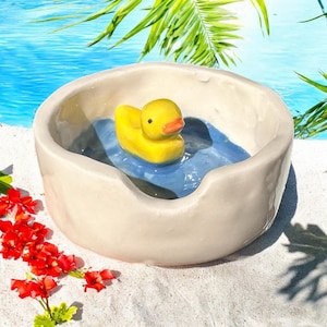 以下が含まれることがあります： 青い水に黄色いゴム製のオモチャの鴨が浮かんでいる、プールのような形をした白いセラミック製のボウル