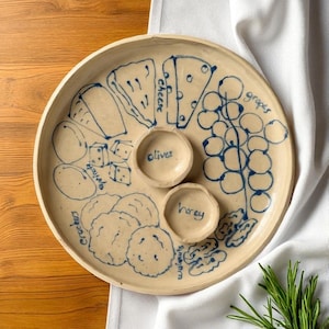 May include: A beige ceramic serving platter with blue illustrations of cheese, grapes, crackers, and walnuts. The platter has two small bowls labeled "olives" and "honey". The platter is on a wooden surface.