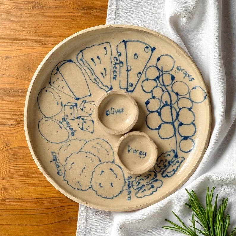 May include: A beige ceramic serving platter with blue illustrations of cheese, grapes, crackers, and walnuts. The platter has two small bowls labeled "olives" and "honey". The platter is on a wooden surface.