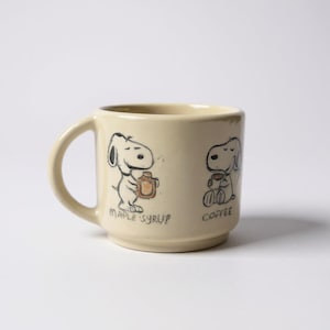 May include: Cream-colored ceramic mug featuring Snoopy illustrations. One Snoopy holds a maple syrup bottle, the other holds a coffee cup. The words "MAPLE SYRUP" and "COFFEE" are printed below the images. The mug has a rounded handle.
