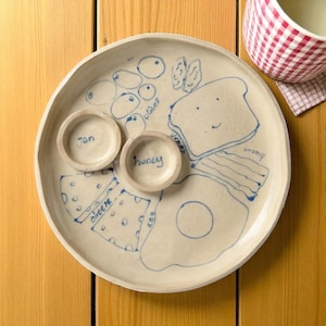 Può includere: Un piatto per la colazione in ceramica dipinto a mano con disegni a tratto blu di alimenti. Include illustrazioni di formaggio, uova, pancetta, toast e olive. Due piccole ciotole sono etichettate "jon" e "honey". Una ciotola a scacchi rossi e bianchi è sullo sfondo.
