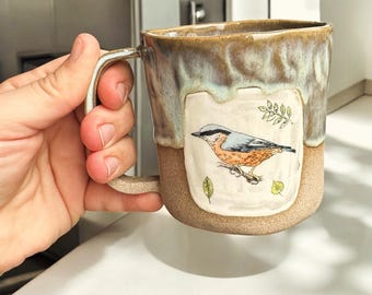 May include: A ceramic mug with a light brown glaze and a white design of a bird with green leaves. The mug has a handle and is a light brown color.