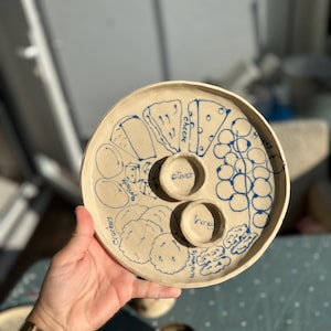 May include: A handmade, off-white ceramic serving platter with blue illustrations of cheese, crackers, olives, honey, walnuts, and grapes. The platter has two small, round wells for olives and honey.