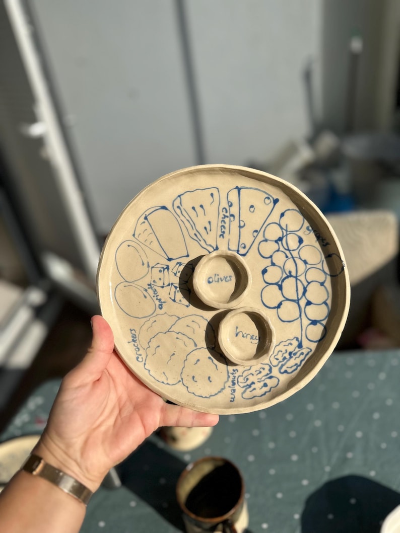 May include: A handmade, off-white ceramic serving platter with blue illustrations of cheese, crackers, olives, honey, walnuts, and grapes. The platter has two small, round wells for olives and honey.