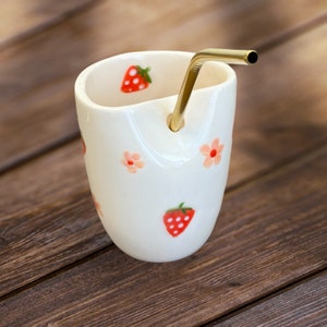 Könnte beinhalten: Weiße Keramiktasse mit einem goldenen Metallstrohhalm, verziert mit einem Erdbeer- und Blumendesign.