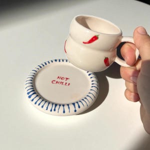 Può includere: Una tazza in ceramica bianca con forma arrotondata e manico, decorata con illustrazioni di peperoncini rossi. È presente anche un sottobicchiere abbinato con bordi a righe blu e bianche e la scritta "HOT CHILLI" in rosso.