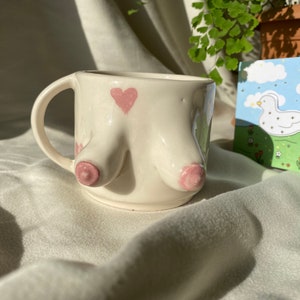 Puede incluir: Taza de cerámica blanca con un corazón rosa y dos protuberancias rosadas en relieve. La taza está sobre una superficie de tela blanca.