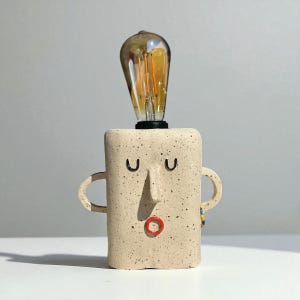 Può includere: Lampada in ceramica beige stravagante con design a volto. La lampada ha un corpo rettangolare con naso, occhi chiusi e bocca rossa. Presenta due manici e una lampadina in stile vintage.