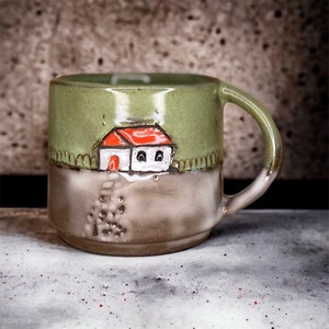 Puede incluir: Una taza de cerámica verde con un esmalte marrón y una imagen pintada a mano de una casa blanca con un techo rojo. La casa está rodeada de hierba verde y una valla.
