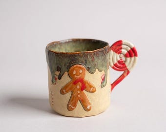 May include: A handmade ceramic mug with a gingerbread man design. The mug features a cream-colored base with a brown gingerbread man wearing a red scarf. The handle is red and white striped, resembling a candy cane. The rim has a green and brown glaze.