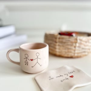 Op de afbeelding: Een witte keramische mok met een handgetekend ontwerp van twee stokpoppetjes die een hart vasthouden. De mok staat op een witte keramische onderzetter met de tekst "You make my heart smile" en een klein rood hart.