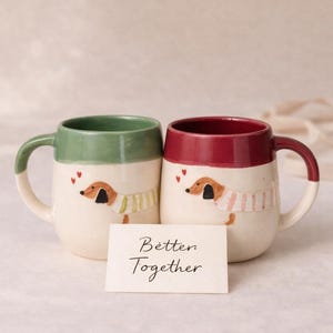 Puede incluir: Dos tazas de cerámica con ilustraciones de perros salchicha. Una taza tiene un borde y asa verdes, la otra tiene un borde y asa burdeos. Ambas tazas tienen una base color crema y una tarjeta con el texto "Better Together".