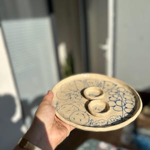 May include: A handmade ceramic serving platter with a light beige base and blue illustrations. The platter features two small, round bowls and is decorated with drawings of olives, nuts, and crackers. The word "crackers" is visible on the platter.