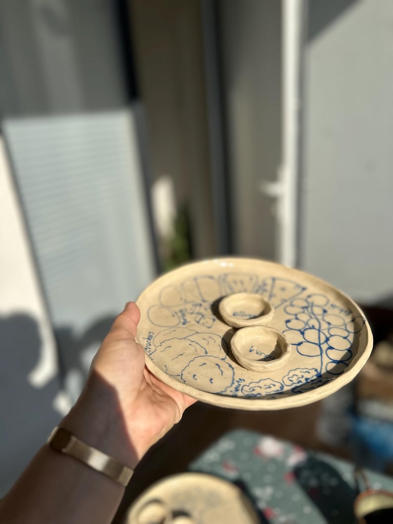 May include: A handmade ceramic serving platter with a light beige base and blue illustrations. The platter features two small, round bowls and is decorated with drawings of olives, nuts, and crackers. The word "crackers" is visible on the platter.