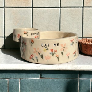 May include: A white ceramic serving dish with a smaller dip bowl attached. The dish is decorated with a floral pattern and the words "Dip Me" and "Eat Me".