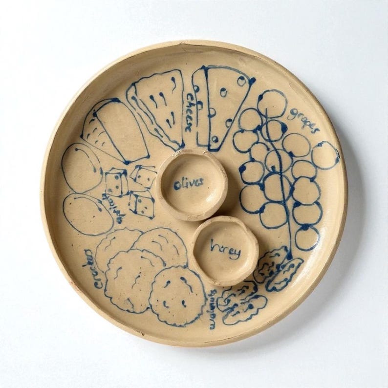 May include: A beige ceramic serving platter with blue illustrations of cheese, grapes, crackers, olives, honey, and walnuts. The platter has two small bowls for olives and honey. The word "cheese" is written on the platter.
