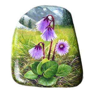 Può includere: Una pietra dipinta con un'illustrazione dettagliata di fiori viola con foglie e steli verdi. Lo sfondo raffigura un campo erboso, montagne e un cielo nuvoloso. La pietra ha una finitura liscia e lucida.