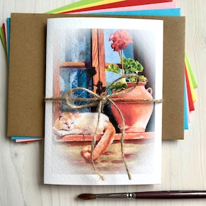 May include: A watercolor greeting card featuring a sleeping orange and white cat on a windowsill, with a terracotta pot holding a red flower. The card is tied with twine and comes with a stack of colorful envelopes and a brown envelope.