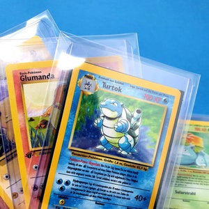 Può includere: Diverse carte collezionabili Pokémon sono visibili, racchiuse in custodie protettive trasparenti. Le carte presentano illustrazioni colorate di vari personaggi Pokémon, con testo in più lingue. Lo sfondo è blu brillante.